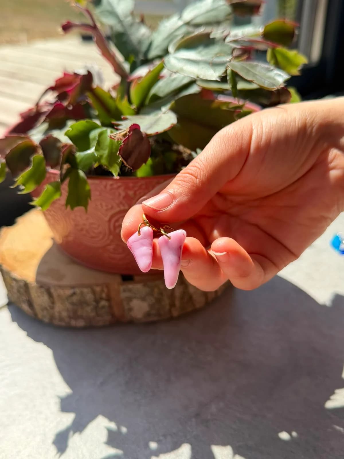 Main tenant une paire de boucles d'oreilles pendantes en verre marbré rose devant une plante en pot.
