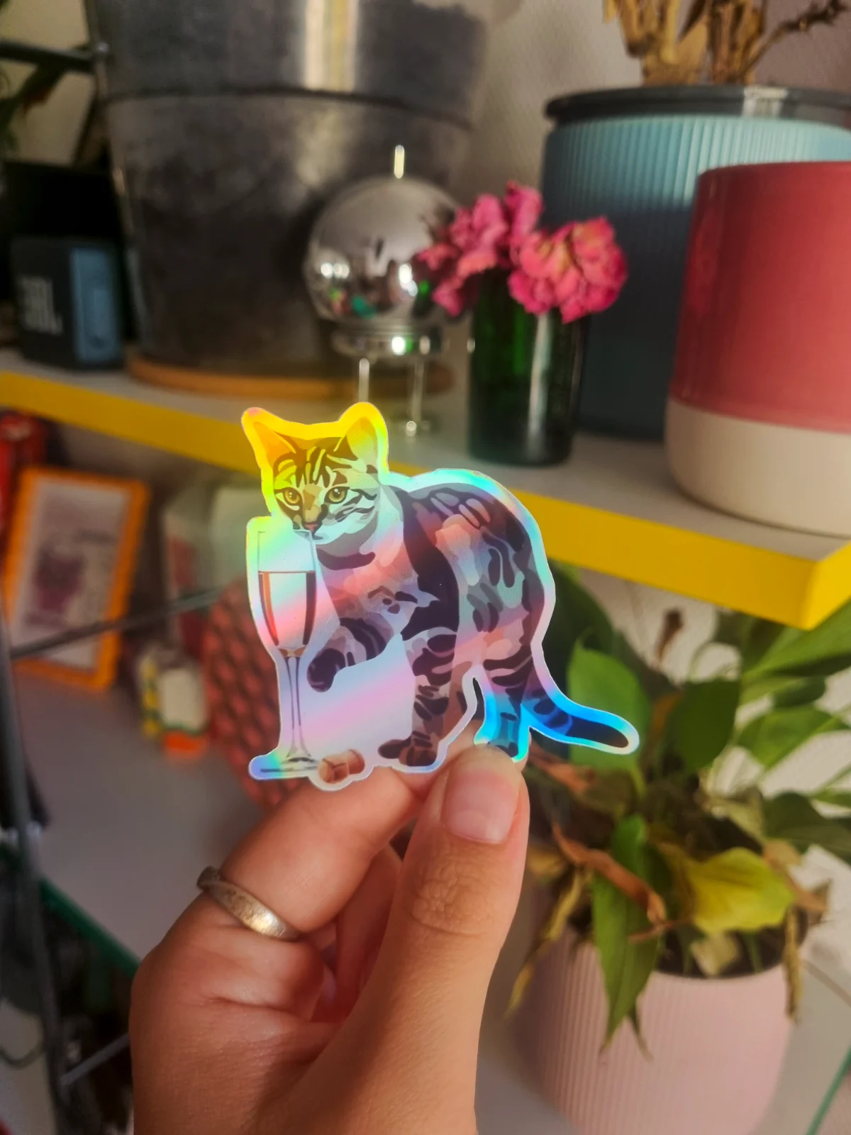 Sticker holographique illustrant un chat tigré près d'une flûte à pied et d'un bouchon, tenu en main.