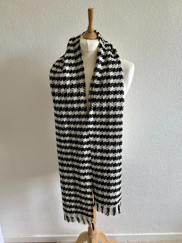 Écharpe à motifs tissés noirs et blancs avec franges, portée sur un buste de mannequin blanc.