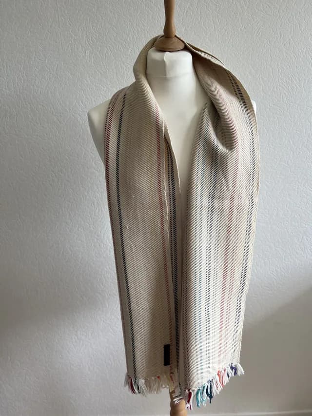 Écharpe tissée couleur champagne à rayures colorées et franges, disposée sur un buste de mannequin blanc.