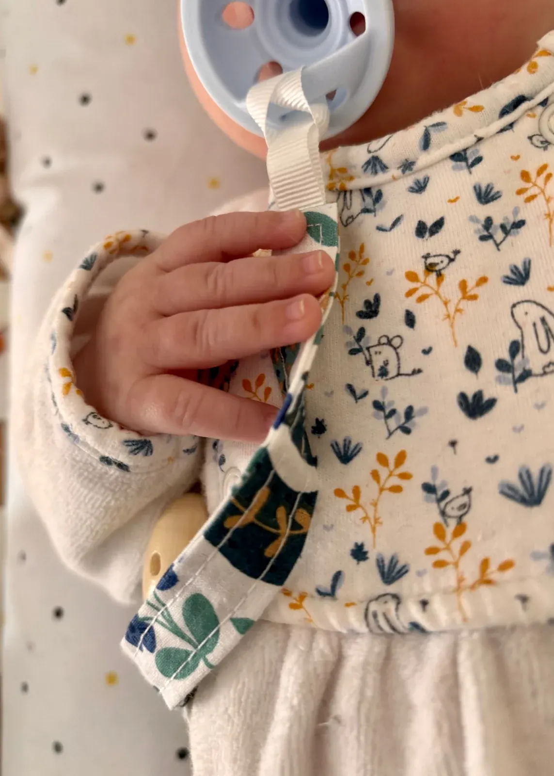 Attache-tétine en tissu à motifs avec clip en bois clair, posée sur un plaid beige