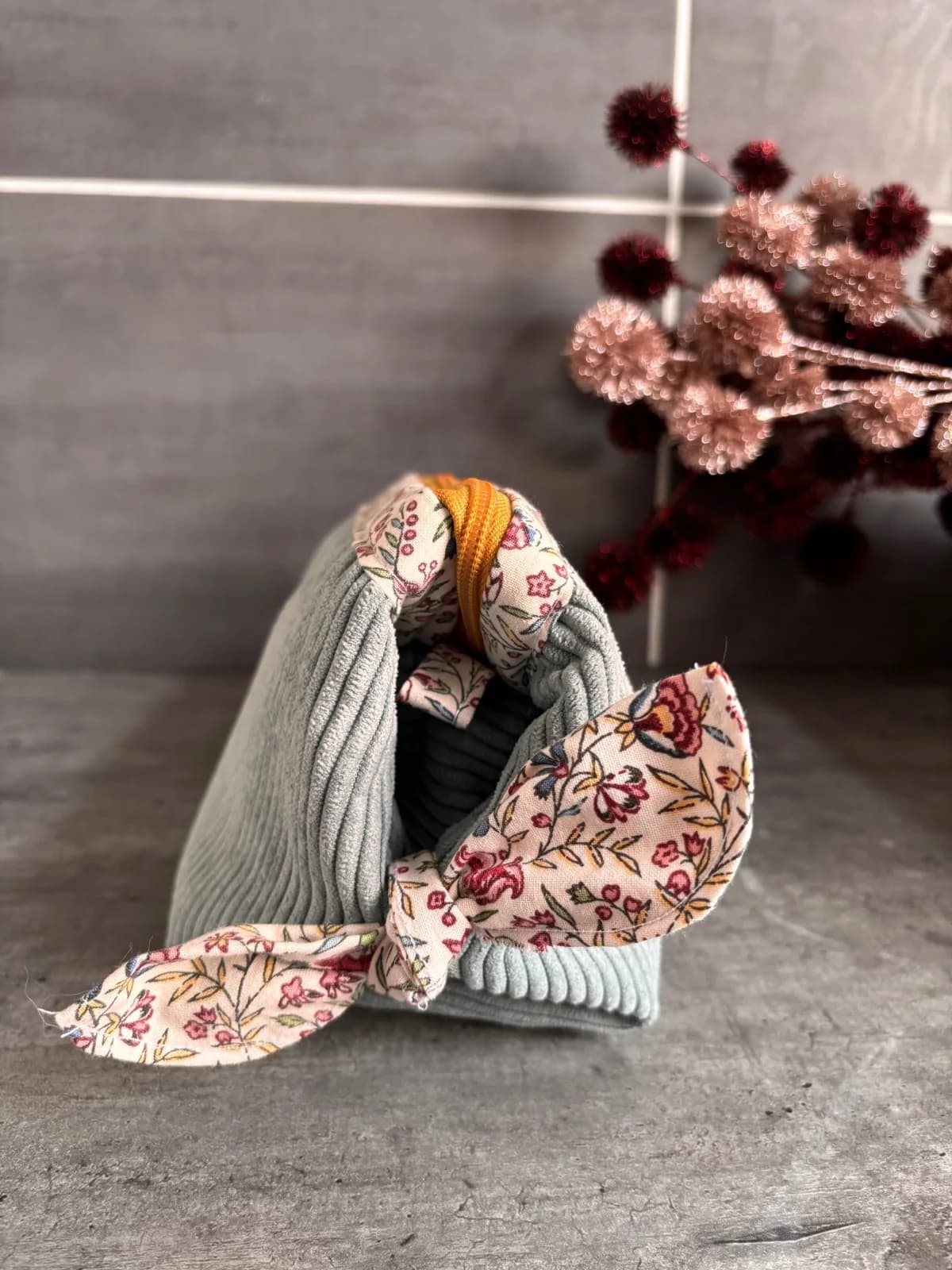 Trousse en velours côtelé vert d'eau avec nœud fleuri sur le côté, posée sur une surface grise.