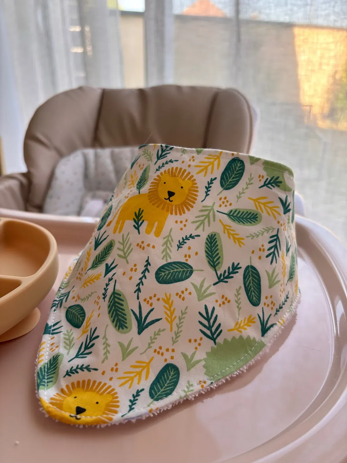 Bavoir bandana blanc à motifs de lion jaune et feuillage vert posé sur une tablette de chaise haute