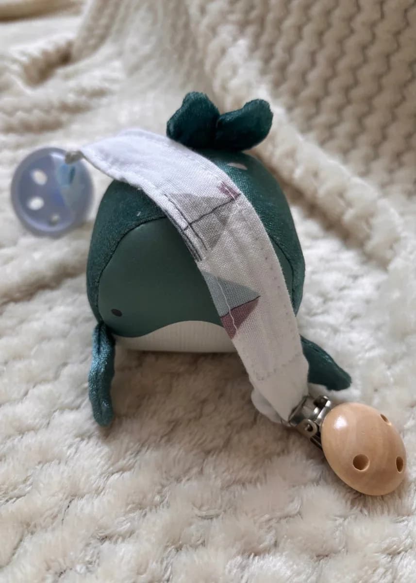 Attache-tétine en tissu à motifs clairs avec clip en bois naturel, posée sur une figurine baleine.