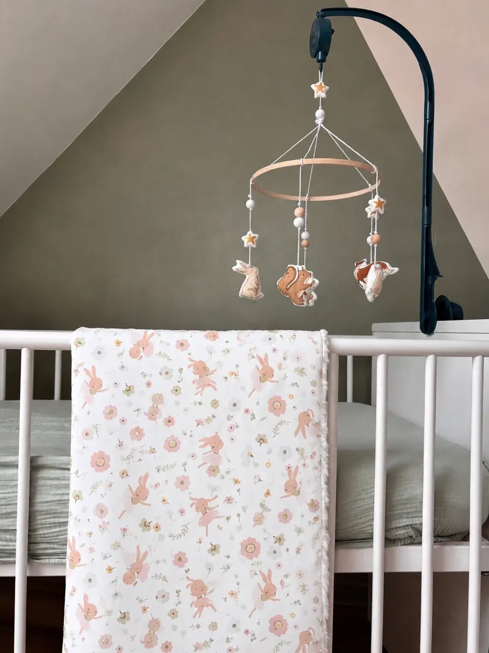 Couverture bébé à motifs de fleurs et lapins posée sur les barreaux d'un lit blanc sous un mobile.