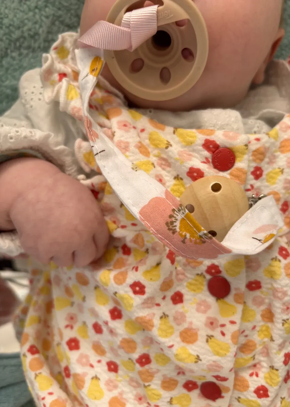 Attache-tétine en tissu à motif floral avec clip en bois portée par un bébé avec sa sucette.