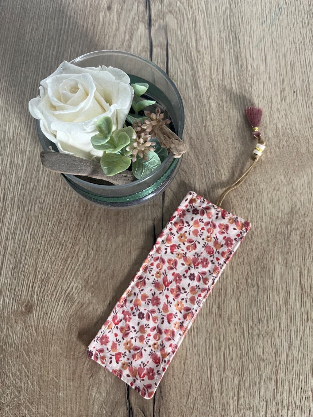 Marque-page textile à petites fleurs roses avec pompon, posé sur une table en bois près d'une rose blanche.