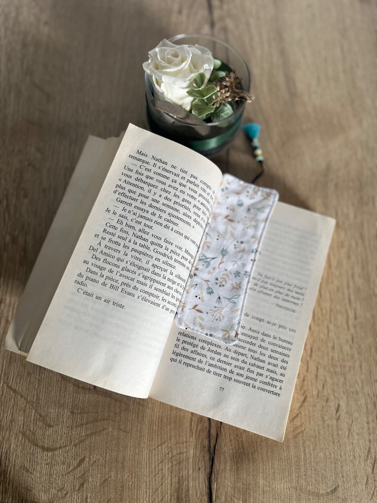 Marque-page en tissu à motifs floraux bleu et beige avec cordon à perles et pompon turquoise sur fond bois.