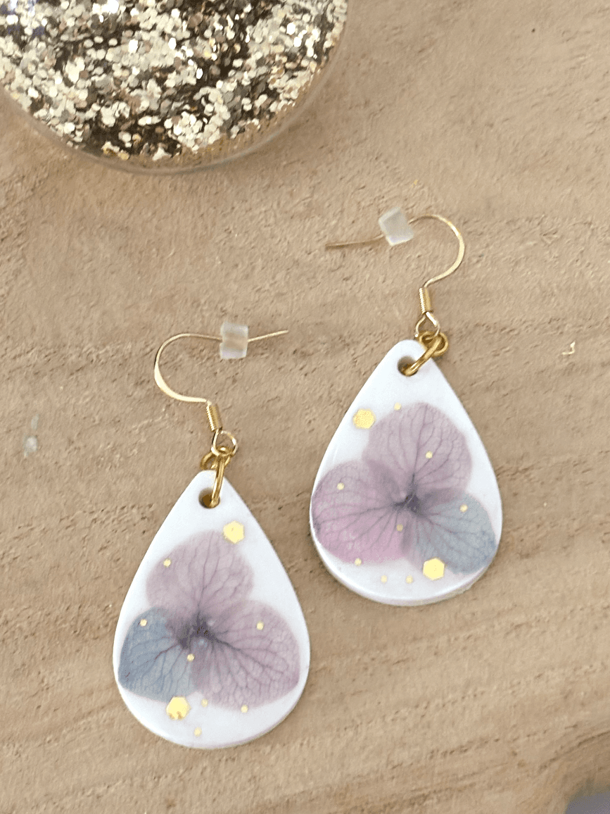 Boucles d'oreilles en goutte blanche et fleur d'hortensia mauve posées à plat sur du bois.