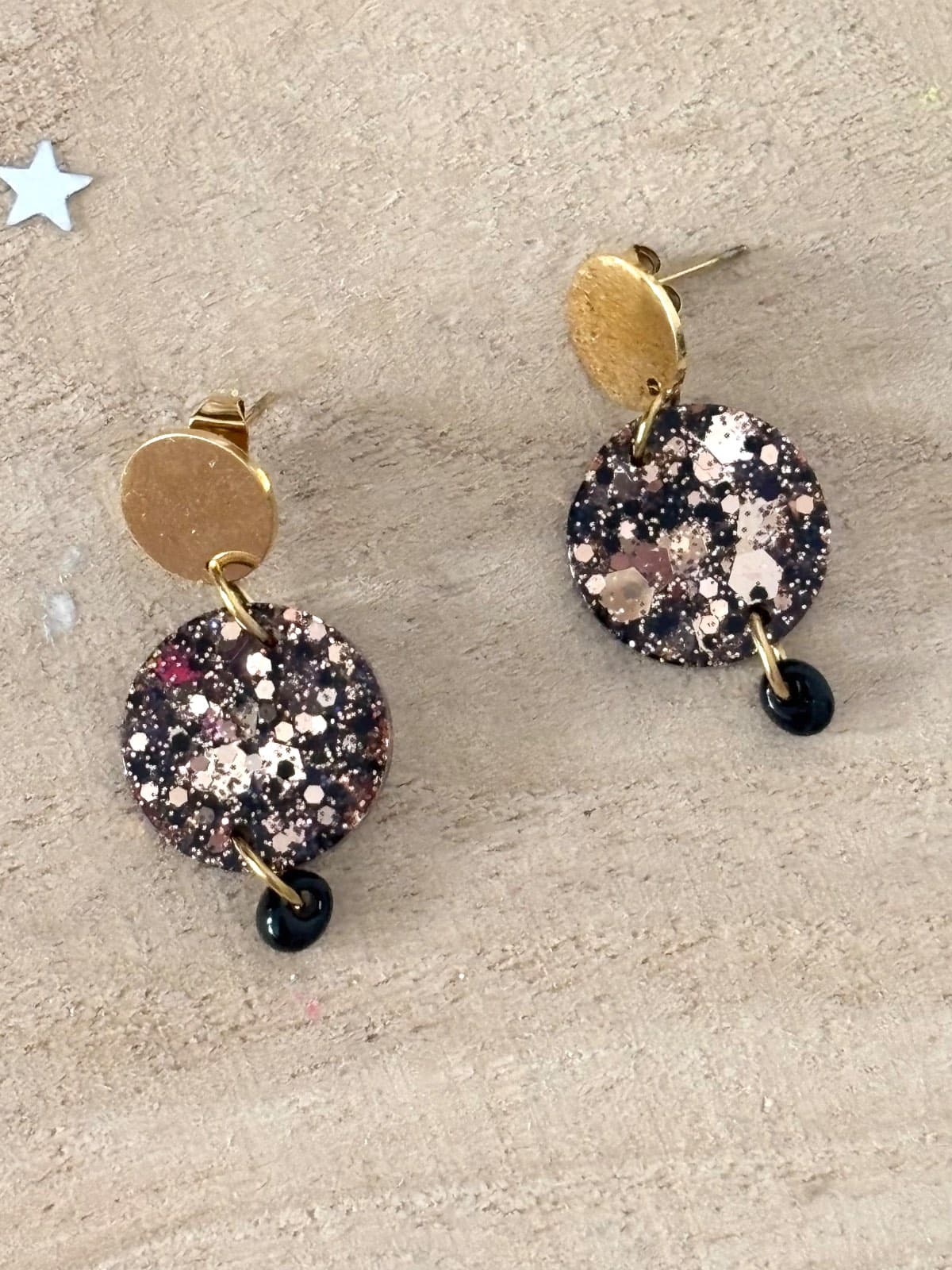 Paire de boucles d'oreilles dorées et médaillons ronds pailletés noir et or posée à plat sur du bois