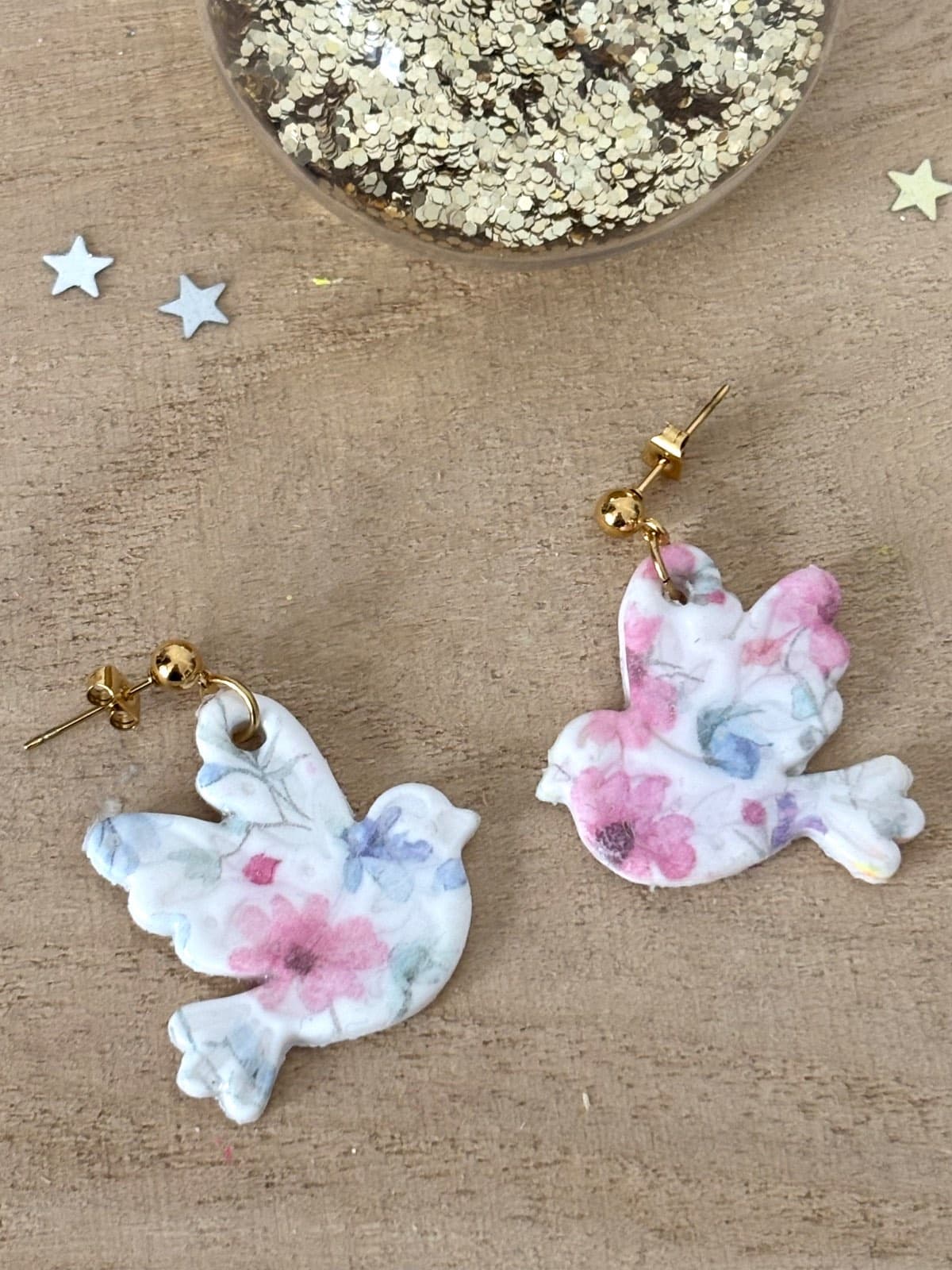 Paire de boucles d'oreilles pendantes en forme d'oiseau à motifs floraux roses et bleus sur fond bois.