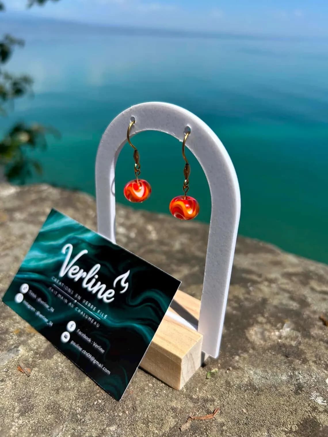 Boucles d'oreilles pendantes en perles de verre marbré orange et rouge sur présentoir blanc face à l'eau.