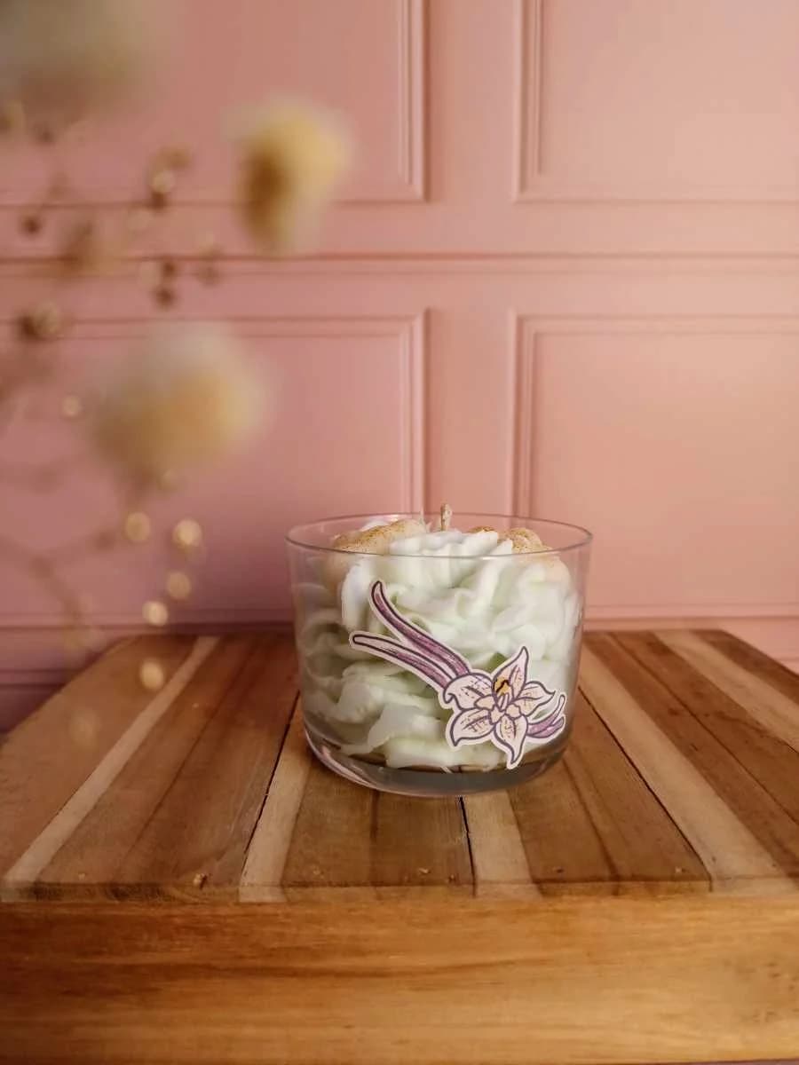 Bougie en verre garnie de chantilly de cire blanche et illustrée d'une fleur de vanille sur un socle en bois.
