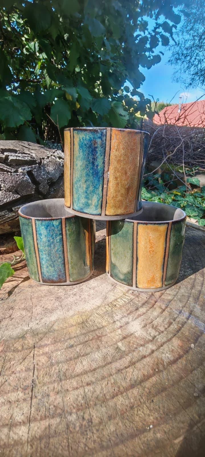 Trois gobelets en céramique à rayures bleu, vert et ocre posés sur une souche de bois.