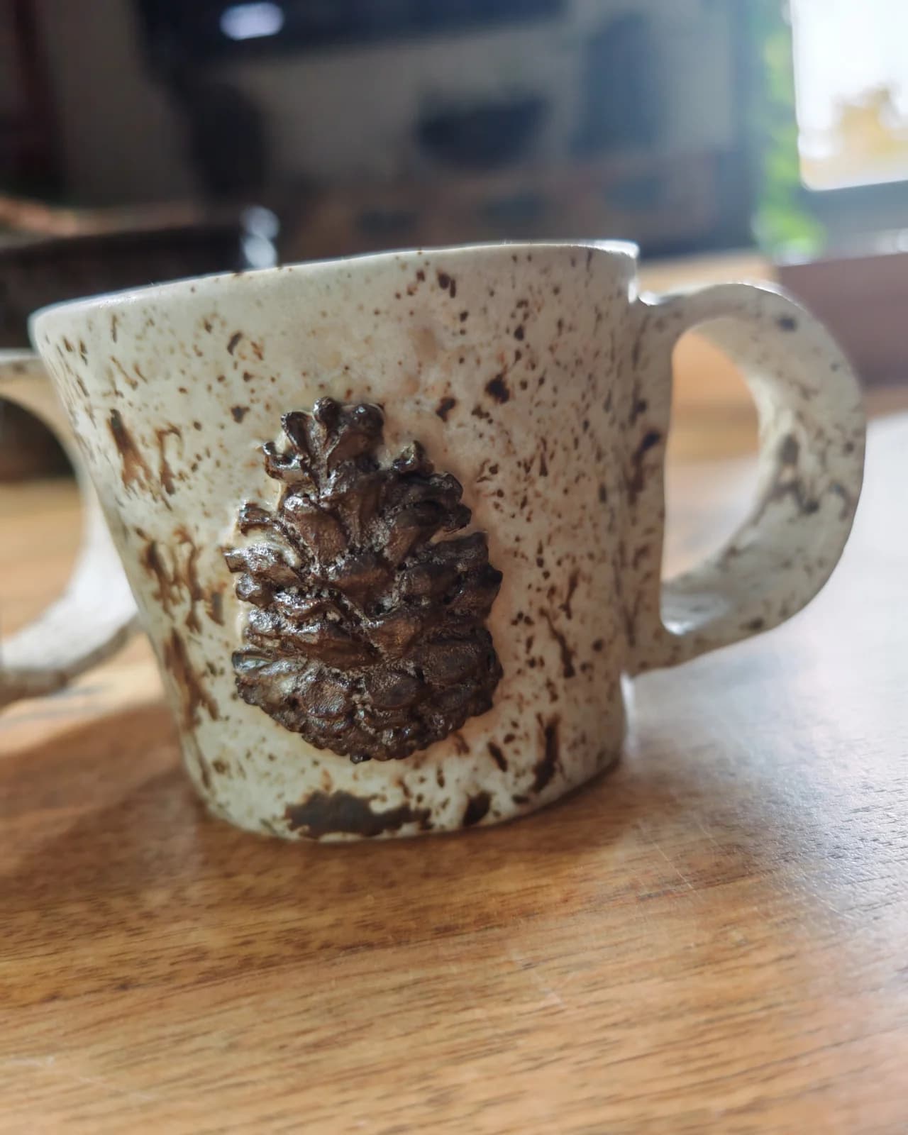 Gros plan sur une tasse artisanale beige mouchetée ornée d'une pomme de pin sculptée en relief.